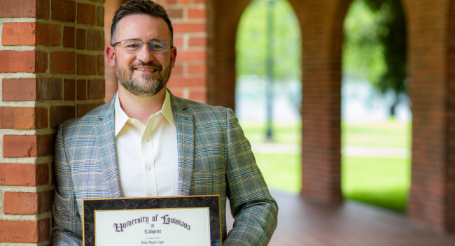 Online MS in Accounting graduate smiles in a white shirt and gray blazer, holding his UL Lafayette master's degree plaque.
